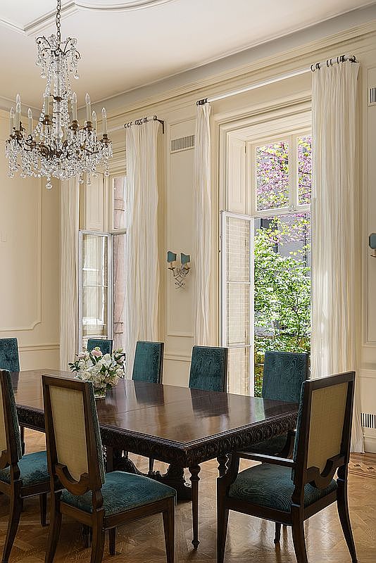 This elegant dining room features a dark wood table surrounded by chairs upholstered in a rich teal fabric. A grand crystal chandelier hangs above the table, while tall windows draped in light-colored curtains allow natural light and views of lush greenery to fill the space. The room is finished with classic wall molding and polished wood flooring, creating a sophisticated and formal atmosphere.