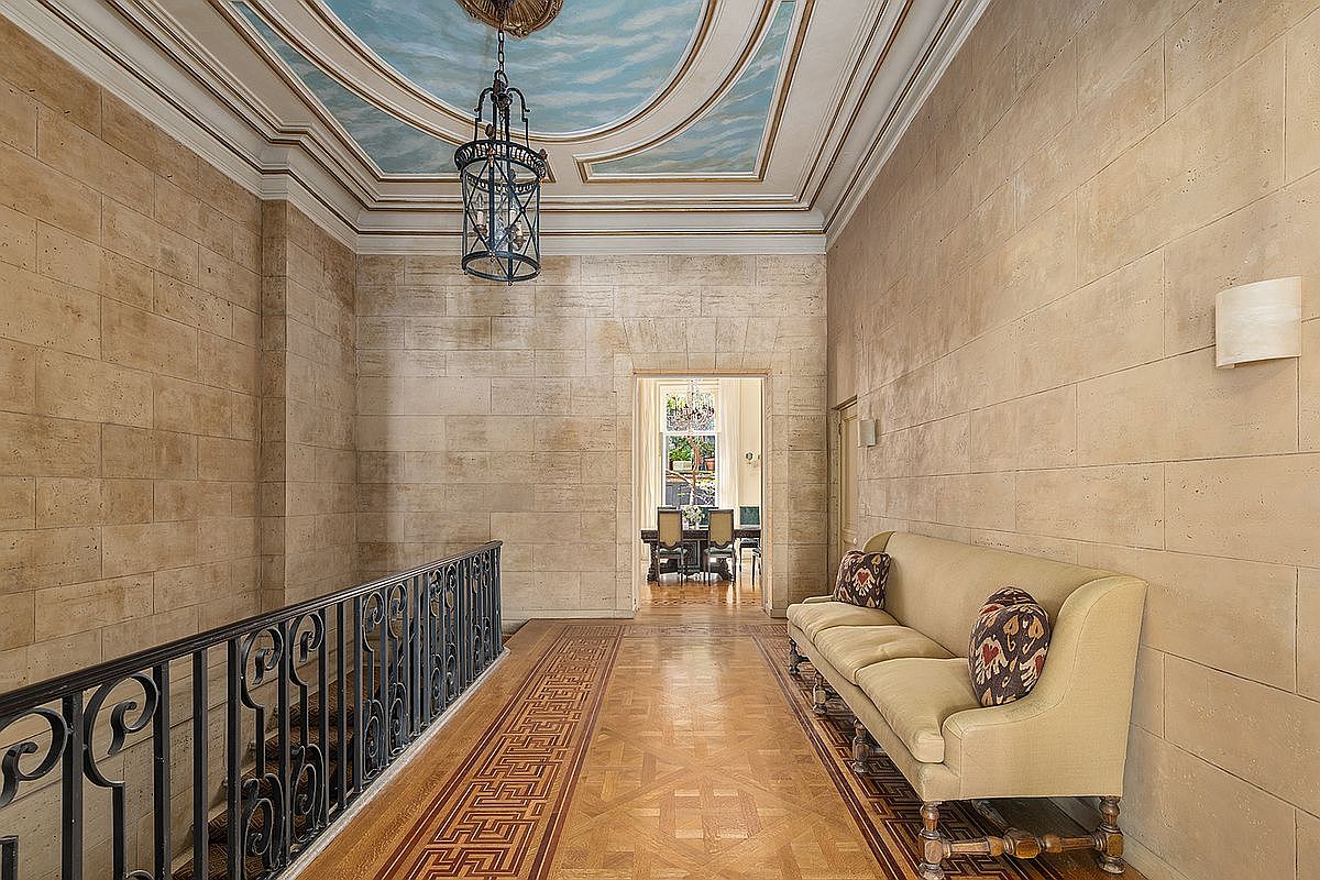 This elegant hallway features stone-textured walls and a sophisticated parquet floor with a decorative border. A wrought-iron railing overlooks the lower level, while a painted ceiling mural depicting a sky adds a dramatic architectural touch. A classic sofa with patterned pillows sits against the wall, leading the eye toward a bright, open doorway that reveals a formal dining area.