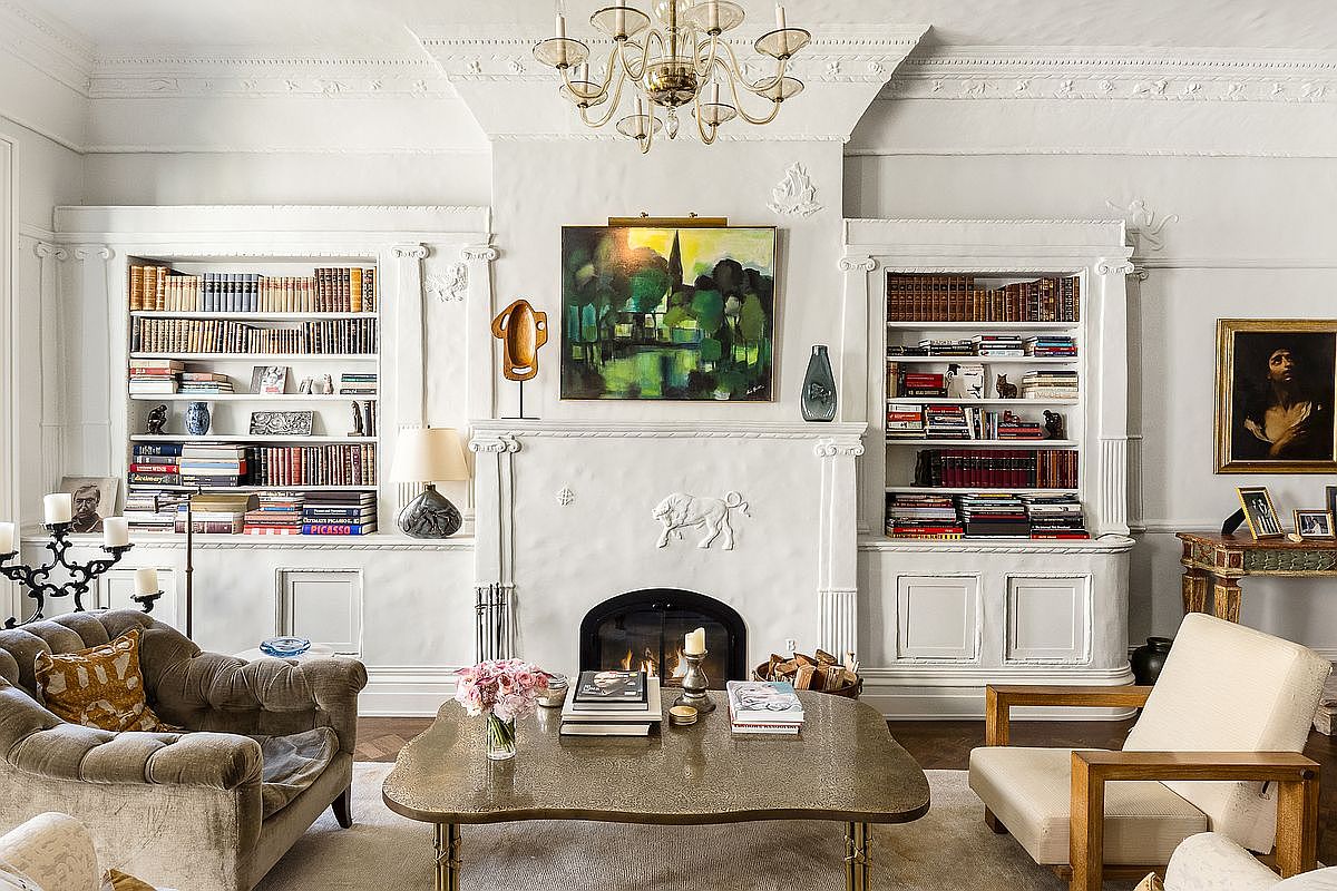 This elegant living room features a symmetrical layout centered around a white fireplace mantel adorned with decorative molding and a landscape painting. Built-in bookshelves flank the fireplace, providing ample storage and display space, while a sophisticated chandelier hangs from the ceiling above a textured coffee table. The room is furnished with a plush, tufted armchair and a modern wooden-framed chair, creating a refined and inviting atmosphere.