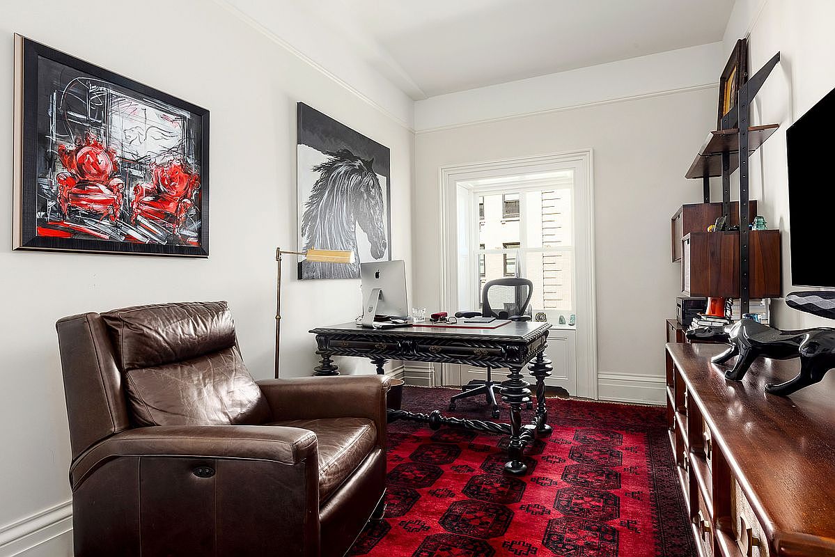 This sophisticated home office features a dark, ornate wooden desk paired with a leather armchair and a vibrant red patterned area rug. The walls are adorned with bold, artistic prints, including a horse portrait and a stylized red chair painting, creating a dramatic and curated aesthetic. A tall shelving unit provides storage, while a window with white shutters allows natural light to brighten the space.