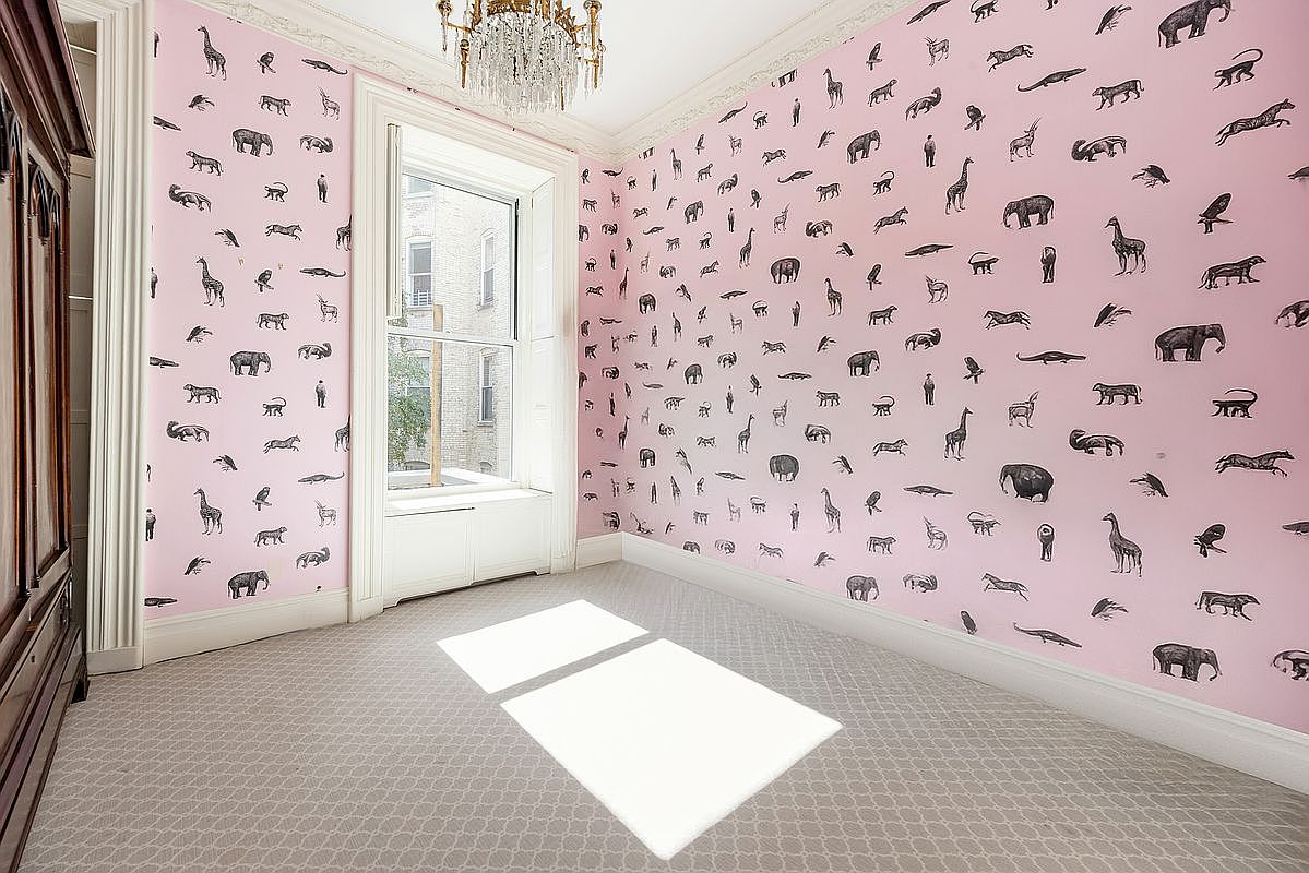 This unique room features whimsical, light pink wallpaper adorned with a repeating pattern of black animal silhouettes, including giraffes, elephants, and crocodiles. A crystal chandelier hangs from the ceiling, while a large window with white trim allows natural light to cast rectangular patterns onto the neutral, patterned carpet. The space is framed by classic architectural molding and a dark wood armoire on the left, creating a playful yet sophisticated atmosphere.