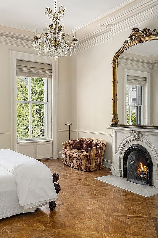 This elegant bedroom features intricate parquet wood flooring and a classic crystal chandelier hanging from the ceiling. A cozy, patterned loveseat sits in the corner near a tall window, while a fireplace with a stone surround and a large, ornate gold-framed mirror serves as a focal point. The room is bathed in natural light, creating a sophisticated and serene atmosphere.