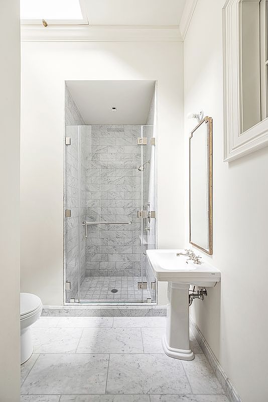 This bright and clean bathroom features a glass-enclosed shower stall with light-toned stone tile walls and flooring. A classic pedestal sink sits to the right, complemented by a tall, framed mirror and neutral wall paint. The space is well-lit and maintains a minimalist, elegant aesthetic.