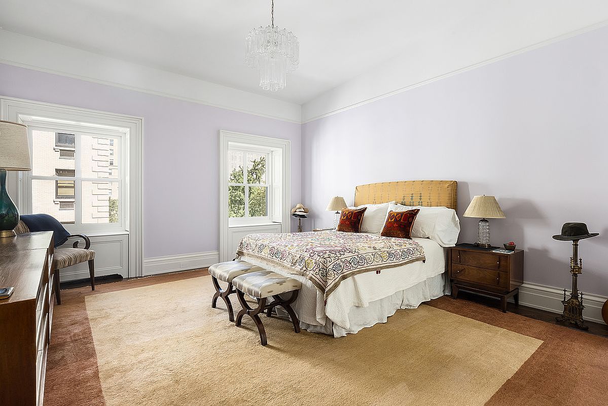 This spacious primary bedroom features soft lavender walls, a large bed with a patterned quilt, and two matching nightstands with lamps. The room is anchored by a large, neutral-toned area rug over dark wood flooring and is illuminated by a decorative crystal chandelier. Two tall windows with white trim provide natural light, creating an airy and elegant atmosphere.