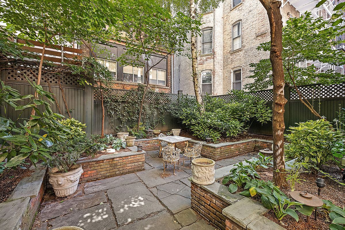 This charming, secluded urban garden features a flagstone patio surrounded by tiered brick planters filled with lush greenery and mature trees. A small bistro set sits in the center, creating an intimate outdoor seating area framed by a dark wooden fence. The space offers a peaceful retreat with a view of the adjacent brick building's exterior, providing a classic city-garden aesthetic.