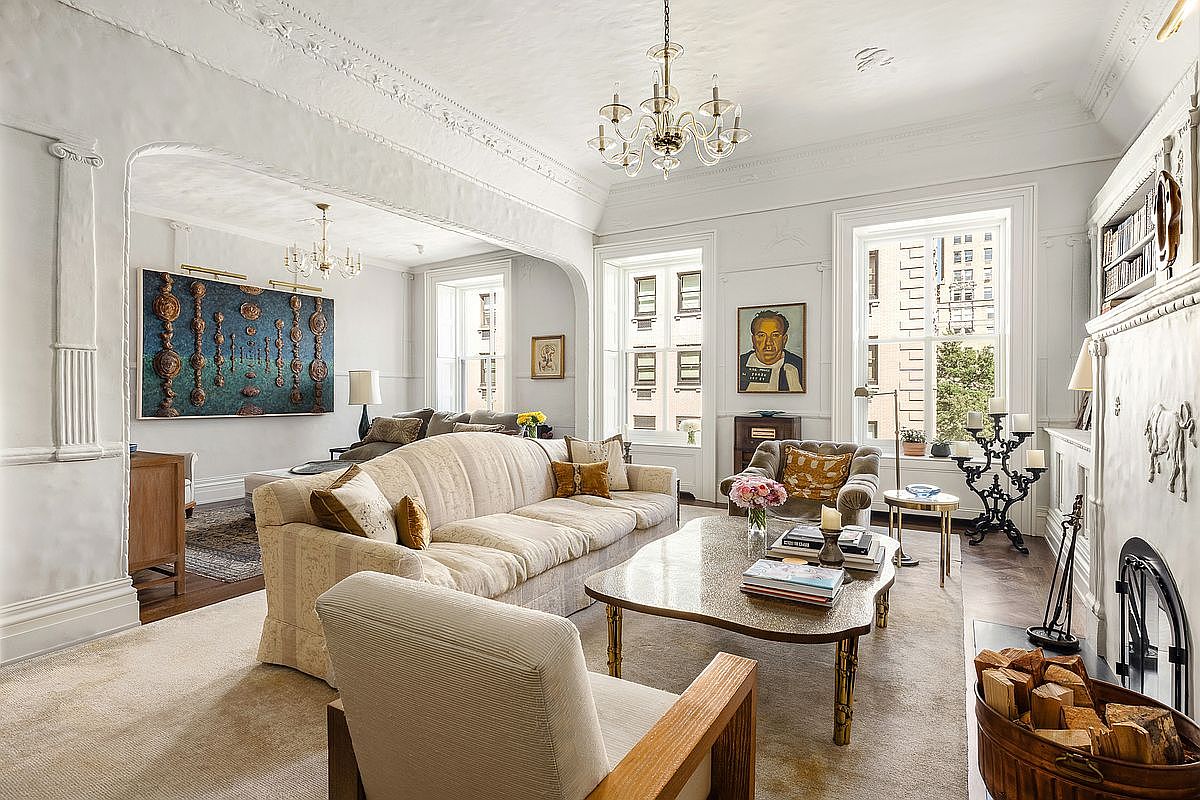 This elegant, light-filled living room features high ceilings with intricate crown molding and a neutral color palette. The space is anchored by a large, plush sofa and a central coffee table, with a fireplace and a wood-filled basket adding warmth to the right side of the room. Large windows allow ample natural light to fill the space, which flows seamlessly into an adjacent sitting area adorned with a prominent textured wall art piece.