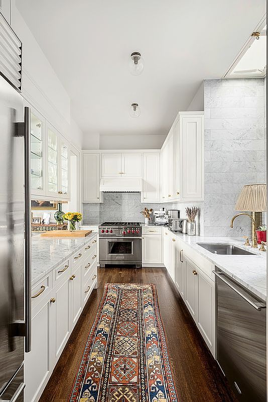 This galley-style kitchen features crisp white cabinetry paired with light-toned stone countertops and a classic subway-tiled backsplash. A vibrant, patterned runner rug adds a pop of color to the dark wood flooring, while stainless steel appliances, including a professional-grade range, provide a modern touch. The perspective looks down the center of the kitchen, highlighting the symmetrical layout and the clean, bright aesthetic.