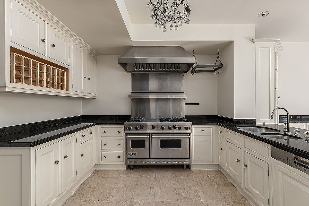 This classic kitchen features white cabinetry with integrated wine storage and dark stone countertops. A professional-grade stainless steel range and matching hood serve as the focal point, complemented by a crystal chandelier overhead. The space is finished with light-toned tile flooring, creating a bright and functional culinary environment.