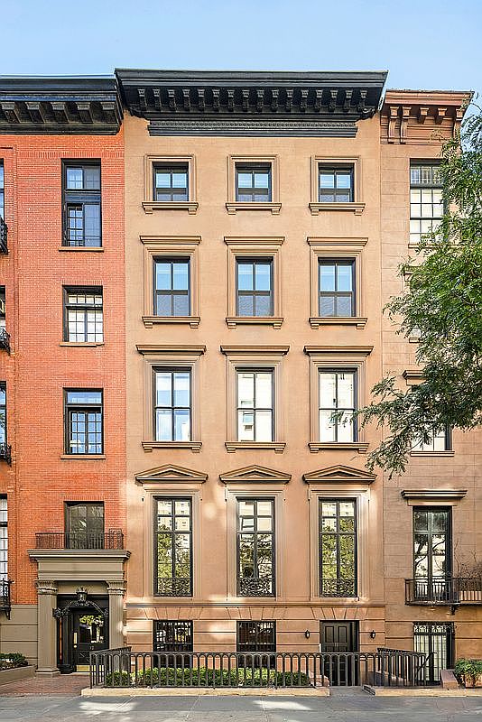 This image captures the front facade of a classic, multi-story brownstone townhouse in an urban setting. The building features a symmetrical design with tall, multi-pane windows, decorative lintels, and a prominent dark cornice at the roofline. A wrought-iron fence encloses a small front garden area, leading to a recessed entryway with a detailed door surround.