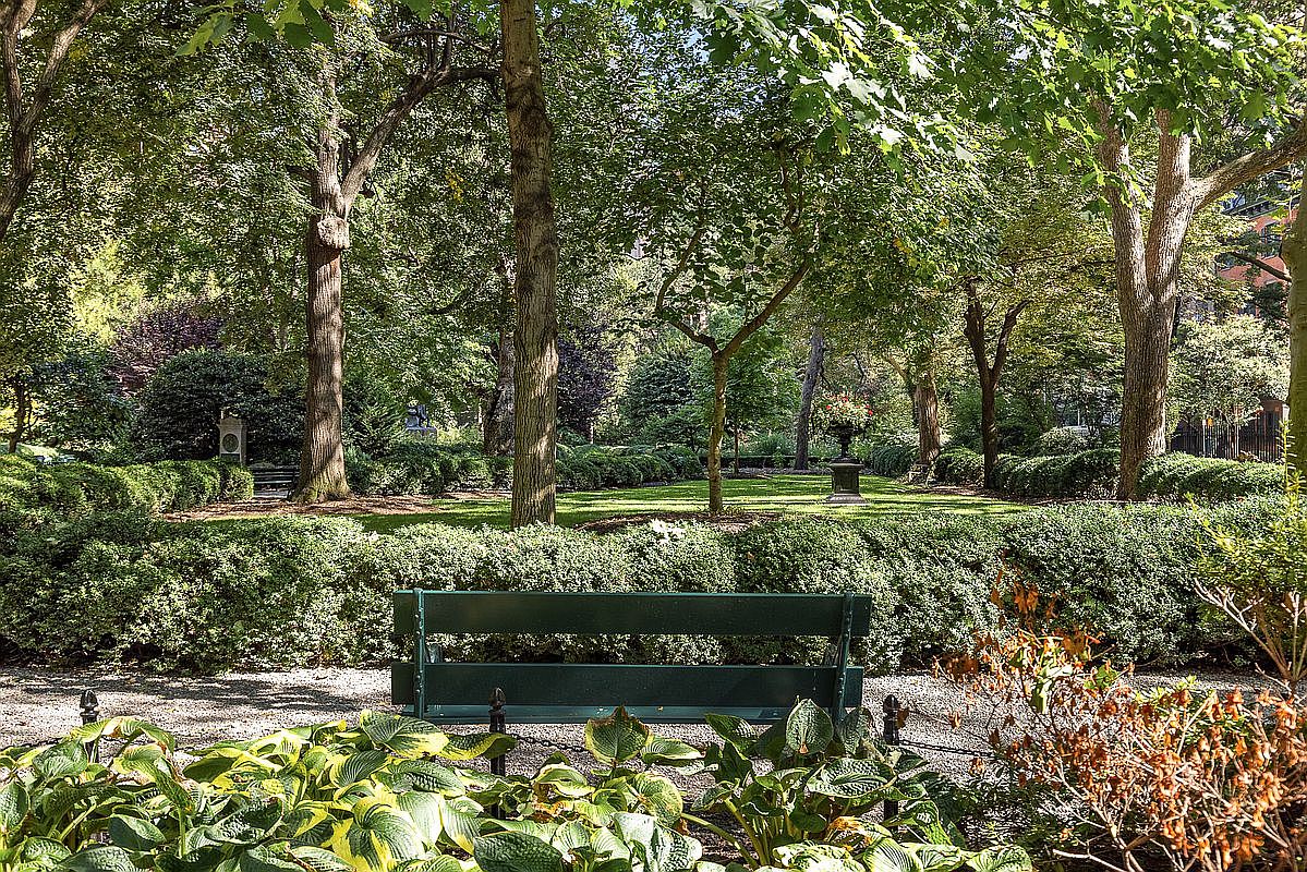 This serene park setting features a lush, manicured lawn framed by mature trees and neatly trimmed hedges. A dark green bench sits in the foreground on a gravel path, providing a peaceful vantage point overlooking the garden. The scene is bathed in dappled sunlight, creating a tranquil and inviting atmosphere typical of a well-maintained community green space.