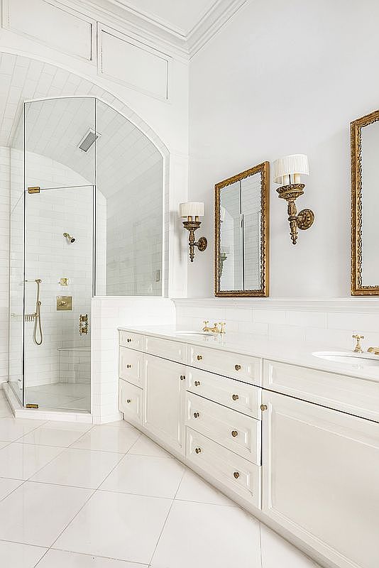 This bright and elegant primary bathroom features a double vanity with white cabinetry, gold-toned hardware, and matching wall sconces flanking ornate mirrors. To the left, a glass-enclosed shower with white subway tile walls and a built-in bench creates a clean, airy aesthetic. The space is finished with large, light-colored floor tiles, offering a sophisticated and cohesive design.
