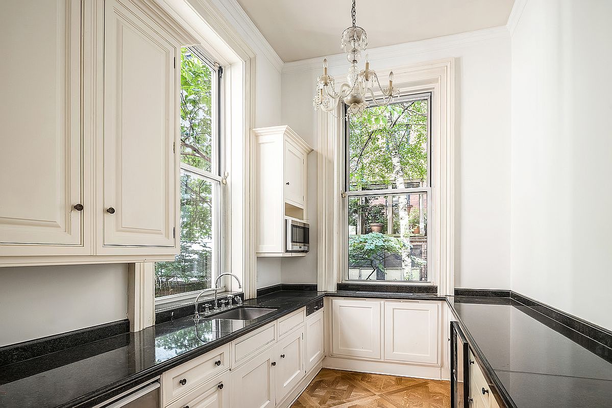 This elegant galley-style kitchen features classic white cabinetry paired with dark stone countertops and a polished finish. The space is brightened by two tall windows that provide a view of lush greenery outside, while a decorative crystal chandelier adds a touch of sophistication above the workspace. The room is finished with warm-toned patterned wood flooring, creating a refined and inviting atmosphere.