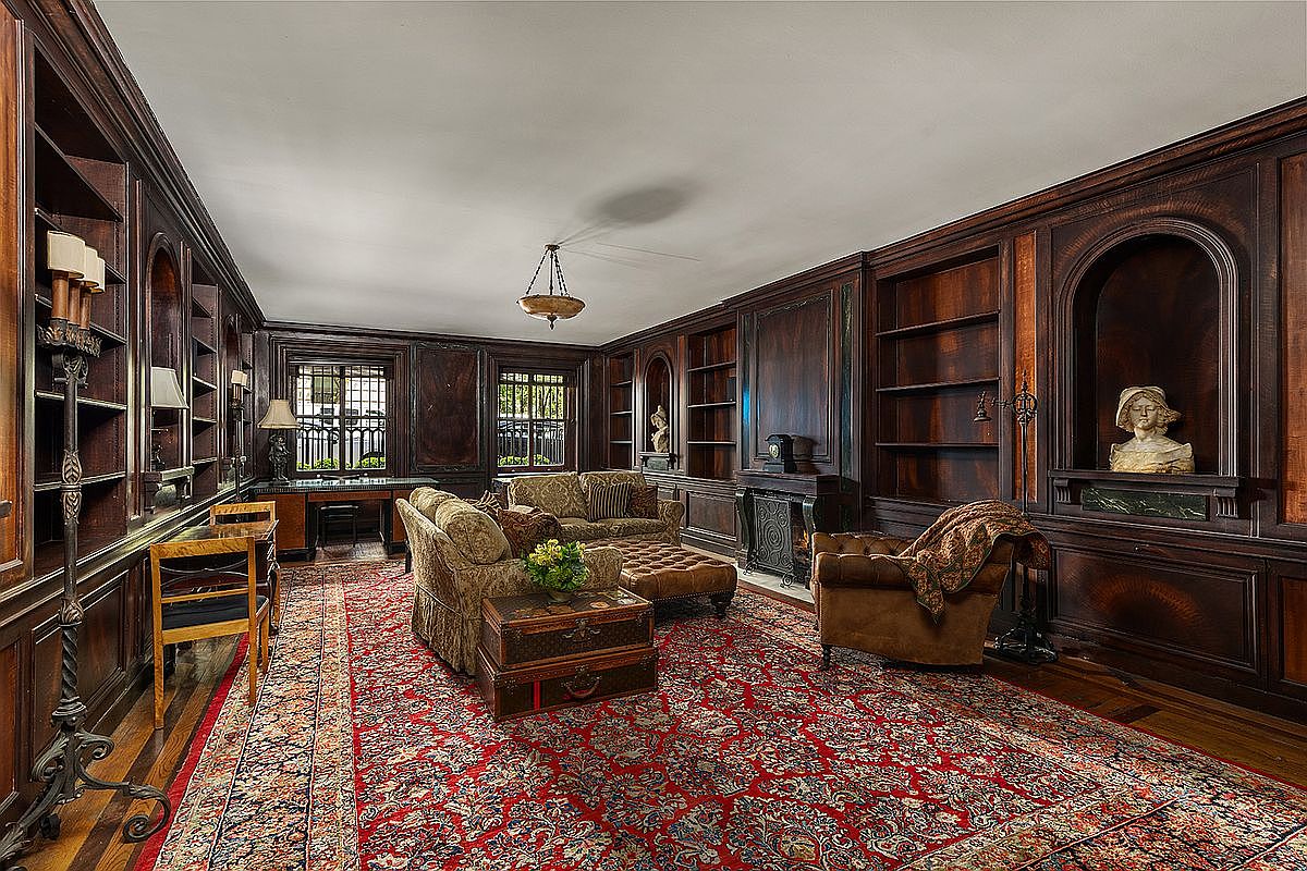 This sophisticated study features rich, dark wood paneling throughout, creating a classic and scholarly atmosphere. The room is furnished with a patterned sofa, a leather armchair, and a desk positioned near the windows, all set atop a large, ornate red area rug. A fireplace with a dark surround serves as a focal point, while built-in bookshelves and arched alcoves displaying decorative busts add to the room's traditional, library-like aesthetic.