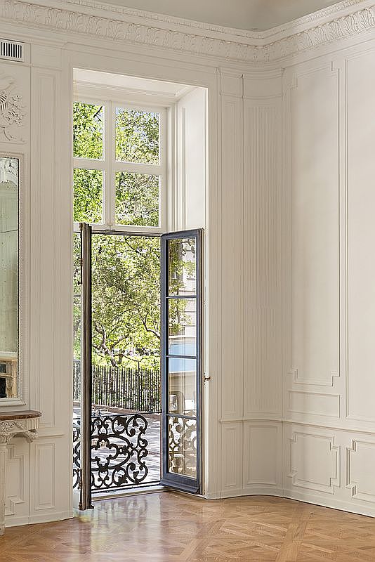 This elegant room features classic architectural details, including intricate crown molding and traditional wall paneling painted in a crisp, neutral tone. A tall, multi-paned glass door opens onto a small balcony with ornate iron railings, allowing natural light to flood the space and providing a view of lush green trees outside. The room is finished with warm-toned, patterned wood flooring, creating a sophisticated and timeless atmosphere.