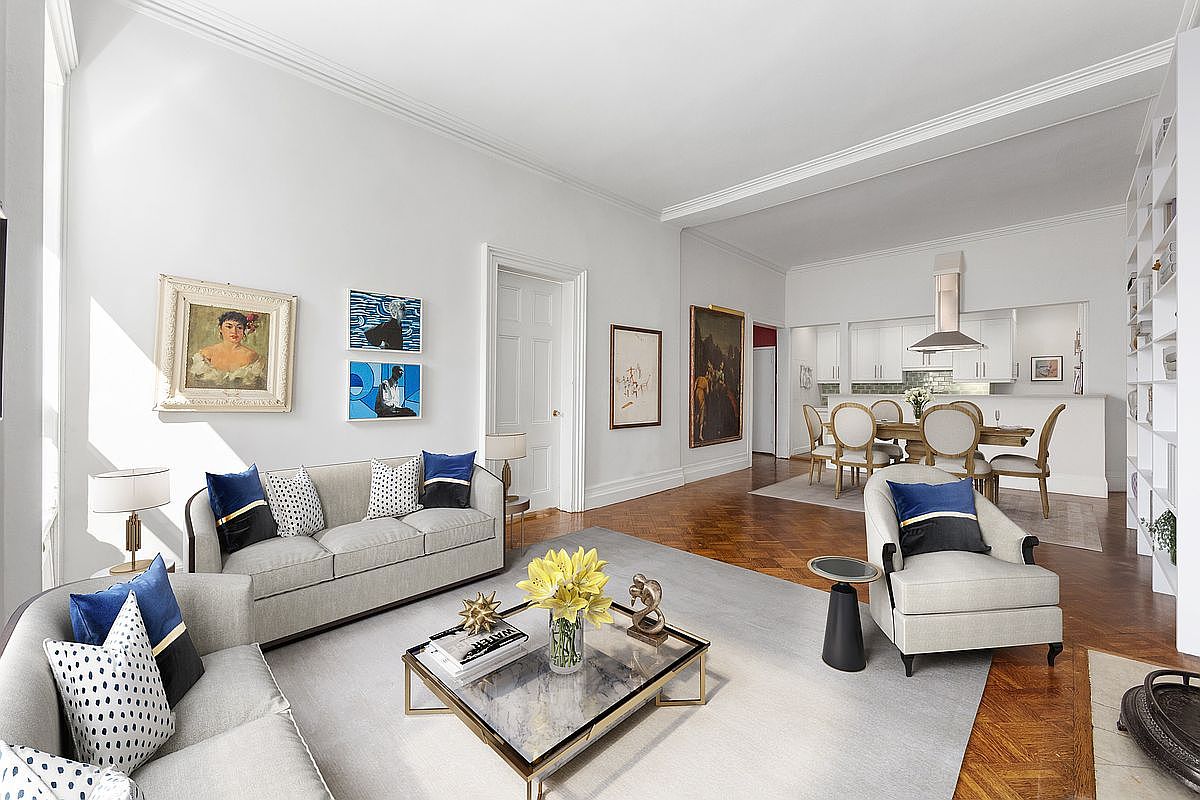 This bright and airy living room features high ceilings, polished herringbone wood floors, and a neutral color palette accented by deep blue throw pillows. The space is furnished with a comfortable sofa and an armchair arranged around a central glass-topped coffee table, while an open-concept dining area with a white kitchen is visible in the background. The room is decorated with various framed artworks and built-in shelving, creating a sophisticated and inviting atmosphere.