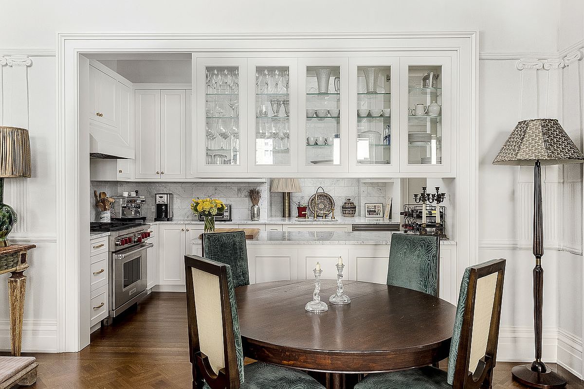 This elegant kitchen and dining area features a classic, sophisticated design with white cabinetry and glass-fronted upper cabinets. A round dark wood dining table sits in the foreground, surrounded by chairs with green velvet upholstery, while the kitchen area showcases a professional-grade range and stone countertops. The space is framed by a wide, architectural opening that creates a seamless transition between the dining and cooking zones.