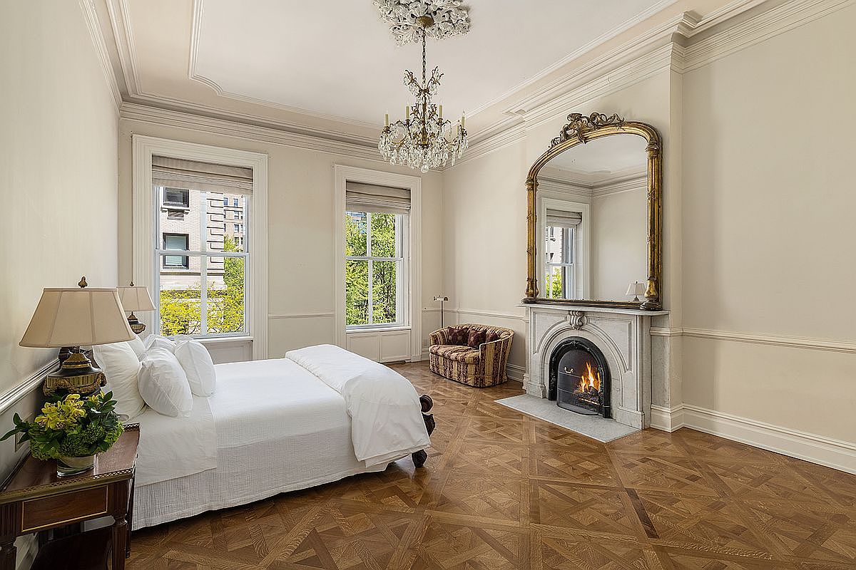 This elegant primary bedroom features intricate parquet wood flooring and a grand, ornate fireplace with a tall, arched mirror above the mantel. A crystal chandelier hangs from the ceiling, casting light over the plush bed and a patterned armchair tucked in the corner. Large windows with neutral shades allow natural light to fill the room, creating a sophisticated and airy atmosphere.