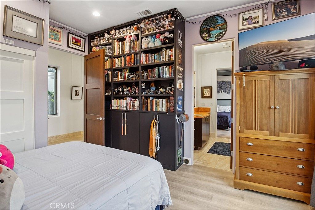 This bedroom features a large, dark-toned built-in shelving unit that serves as a focal point, filled with books and collectibles. To the right, a wooden dresser supports a television, while an open doorway leads into an adjacent bathroom with a vanity and a view of another bedroom. The room is decorated with personal items and wall art, creating a cozy and lived-in atmosphere.