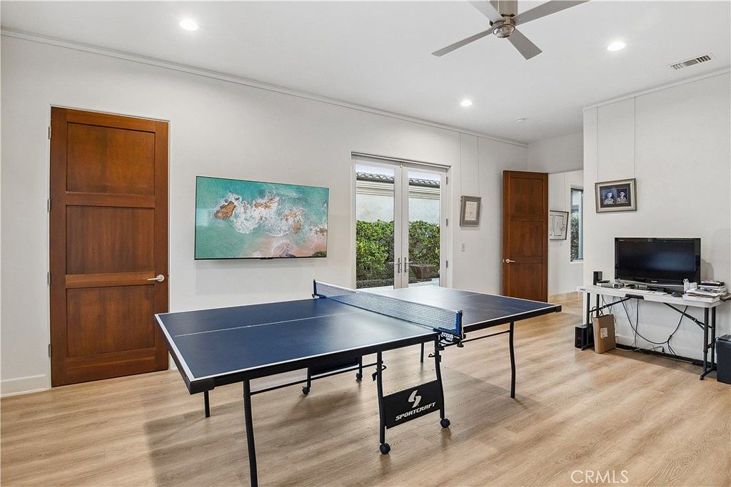This interior space is currently utilized as a game room, featuring a prominent Sportcraft ping pong table as the central focus. The room has light-toned wood-look flooring, white walls, and a ceiling fan, with a set of glass-paned double doors leading to an outdoor area. A television is mounted on a small desk in the corner, and a large landscape-oriented artwork hangs on the main wall, creating a casual and recreational atmosphere.