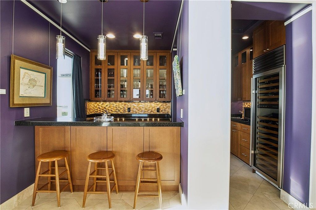 This home bar area features a dark purple color scheme, wood cabinetry, and a black countertop with a mosaic tile backsplash. Three wooden bar stools are positioned at the counter, illuminated by three modern pendant lights hanging from the ceiling. A tall wine refrigerator is built into the adjacent wall, and the space opens into a secondary room with additional cabinetry.