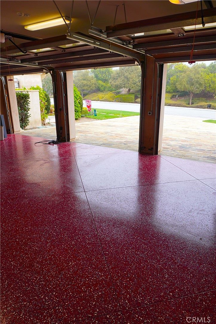This image shows the interior of a spacious, multi-car garage featuring a vibrant, speckled red epoxy floor finish. The open garage doors reveal a paved driveway and a landscaped outdoor area with trees and shrubs under a bright, overcast sky. The perspective is from inside the garage looking out, highlighting the clean, well-maintained condition of the space.