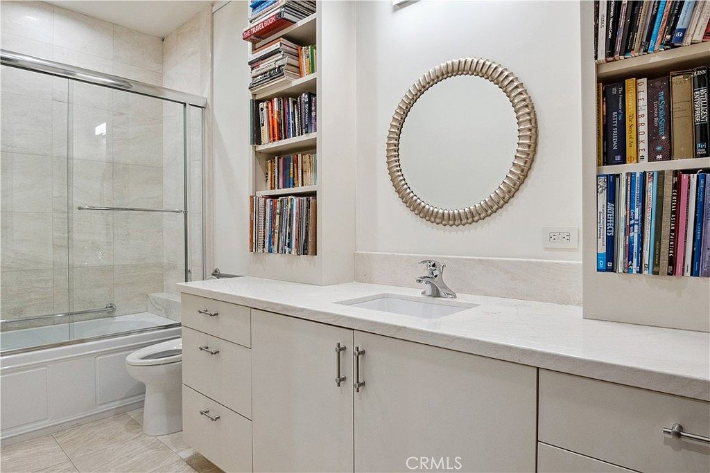 This bathroom features a vanity with a light-colored stone countertop, a white undermount sink, and a circular mirror with a textured metallic frame. Built-in shelving units on either side of the vanity are filled with books, adding a unique, library-like aesthetic to the space. To the left, a glass-enclosed shower and tub combination is visible, featuring light-toned wall tiles and a modern faucet.