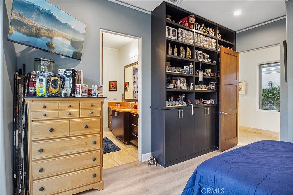 This bedroom features a light wood dresser topped with sports memorabilia and a wall-mounted television, positioned next to a doorway leading into an en-suite bathroom. A large, dark-toned built-in shelving unit provides extensive storage and display space for collectibles, while a wooden door leads to an adjacent area. The room is finished with light-toned flooring and a blue bedspread, creating a personalized and functional living space.