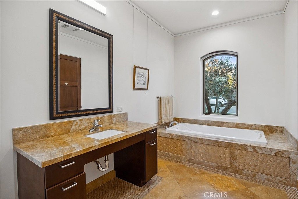This primary bathroom features a vanity with a stone countertop and a dark wood cabinet, paired with a large framed mirror. To the right, a built-in soaking tub is surrounded by matching stone tile work and sits beneath an arched window. The room is finished with large-format stone floor tiles, creating a cohesive and warm aesthetic.