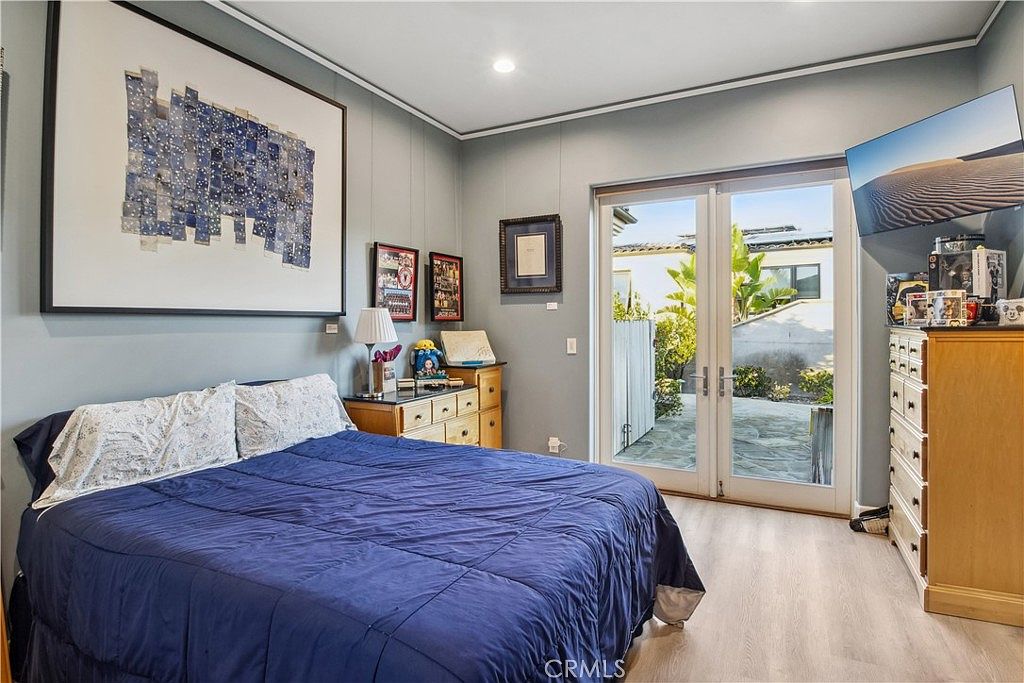 This bedroom features light-toned wood-look flooring and soft blue walls, centered around a bed with a deep blue comforter. A large piece of abstract art hangs above the bed, while a wooden dresser provides storage and surface space for personal items. Large glass doors lead directly to an outdoor patio, allowing for natural light to fill the room.