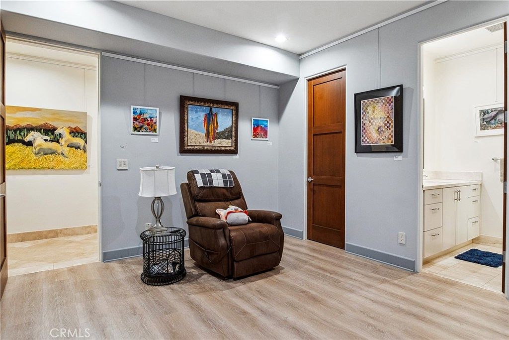 This interior space functions as a cozy sitting area or study, featuring light wood-look flooring and soft gray walls adorned with multiple framed artworks. A brown leather recliner sits beside a small round side table with a lamp, while a wooden door leads to an adjacent room and an open doorway reveals a bright bathroom with a vanity. The room has a clean, gallery-like aesthetic with recessed lighting and a neutral color palette.