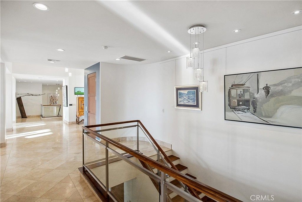 This interior view captures a bright, open-concept landing area featuring a staircase with a glass and wood railing. The space is finished with light-colored tile flooring and white walls, adorned with framed artwork and a modern multi-pendant light fixture. The perspective looks down from the landing toward a hallway, emphasizing the clean, contemporary architectural lines of the home.