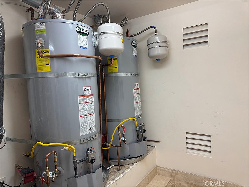 This utility space features two large A.O. Smith water heaters positioned side-by-side, equipped with expansion tanks and copper plumbing connections. The setup includes a red circulation pump attached to the base of the left unit, with yellow gas lines and various valves visible throughout the installation. The area is finished with tiled flooring and two wall-mounted ventilation grilles, indicating a dedicated mechanical or utility closet.