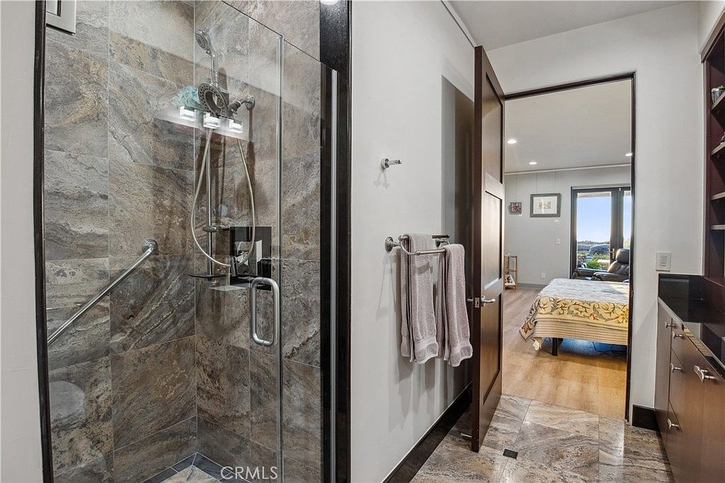 This bathroom features a walk-in shower with textured stone-look wall tiles and a glass enclosure, equipped with a handheld showerhead and a grab bar. The space transitions into a bedroom area through an open doorway, showcasing a neutral color palette and modern finishes. The perspective captures the functional layout of the bathroom while providing a glimpse into the adjacent living space.