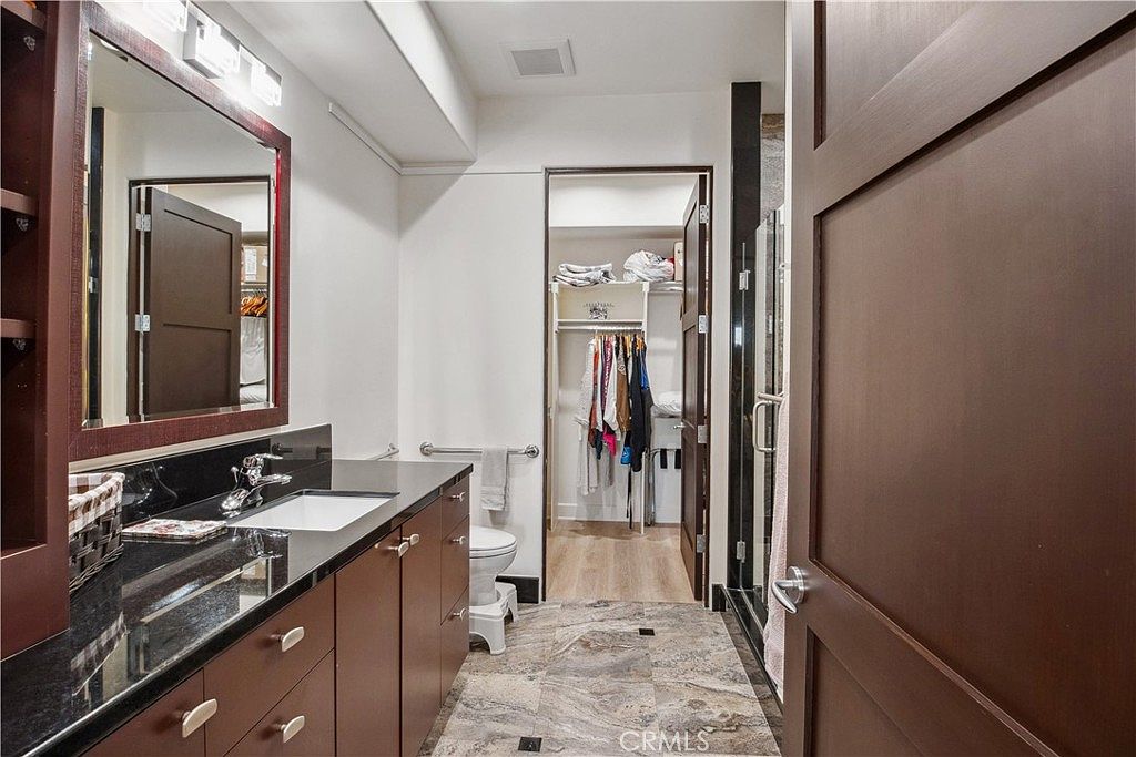 This primary bathroom features a dark wood vanity with a black stone countertop and a rectangular undermount sink. The space includes a toilet, a walk-in closet visible through an open doorway, and a shower area with dark tile accents. The room is finished with patterned stone-look flooring, creating a cohesive and functional layout.