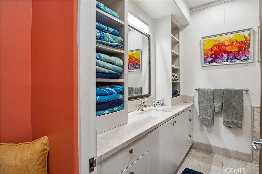 This bathroom features a long vanity with a light-colored stone countertop, a single sink, and ample storage provided by built-in shelving units. The walls are painted a neutral tone, accented by a vibrant, colorful piece of abstract art and a matching reflection in the mirror. The space is finished with light-colored floor tiles and a towel rack holding grey towels, creating a clean and functional aesthetic.