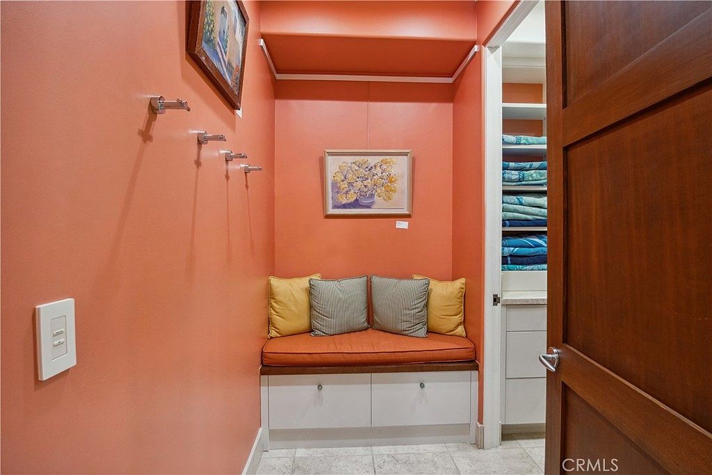 This cozy nook features a built-in bench seat with a vibrant orange cushion and four decorative pillows, set against a matching orange wall. Above the bench, a framed floral artwork adds a touch of color, while a row of wall-mounted hooks provides functional storage. To the right, an open doorway reveals a walk-in closet or storage area with organized shelving, creating a practical and inviting transition space.