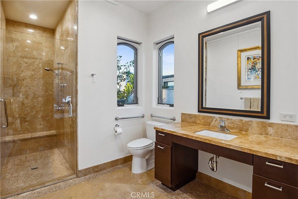 This bathroom features a walk-in shower with stone tile walls and a mosaic floor, adjacent to a vanity with a stone countertop. The space includes a toilet positioned between two tall, arched windows that provide natural light. A large, framed mirror hangs above the vanity, and the room is finished with neutral-toned tile flooring.