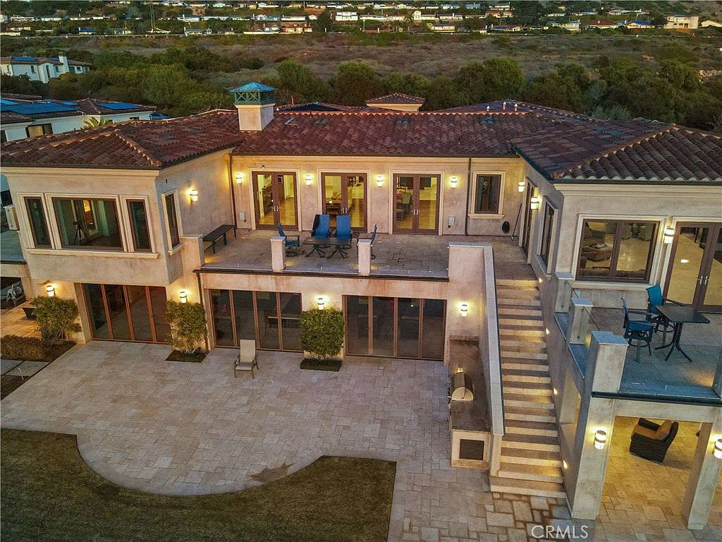 This aerial perspective captures the expansive rear exterior of a Mediterranean-style estate at dusk, highlighting a multi-level layout with a spacious stone-paved patio and an elevated balcony. The home features warm-toned stucco walls, a classic clay tile roof, and numerous glass doors that create a seamless transition between indoor and outdoor living spaces. A wide stone staircase connects the upper balcony to the lower patio, which includes a built-in outdoor kitchen area, all set against a backdrop of rolling hills and a residential neighborhood.