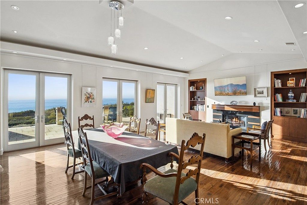 This open-concept living and dining area features polished wood flooring and a bright, airy atmosphere with multiple sets of glass doors opening to an ocean view. The space includes a formal dining table in the foreground and a comfortable seating area centered around a fireplace with a dark, multi-tiered surround. Built-in shelving units flank the fireplace, and a modern pendant light fixture hangs above the dining table, creating a sophisticated and inviting living space.