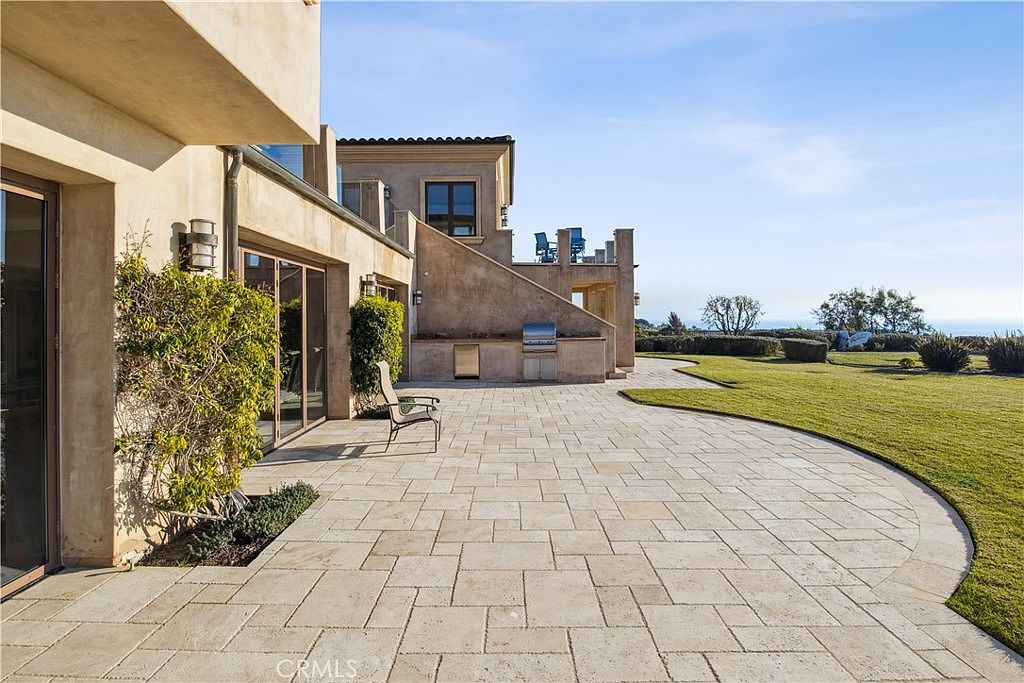 This expansive stone-paved patio offers a seamless transition from the home's interior to a manicured lawn overlooking the ocean. The outdoor space features a built-in grilling station set against a stucco wall, with a single chair positioned to enjoy the coastal view. The architecture showcases a Mediterranean-inspired design with clean lines and a neutral color palette that complements the bright, sunny atmosphere.