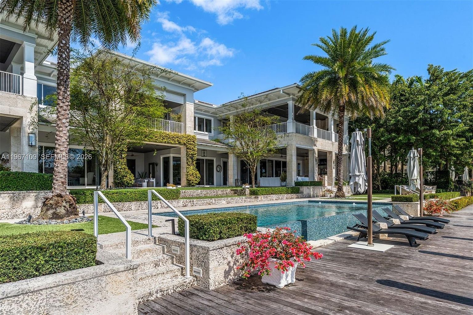 This expansive rear view of a luxury estate showcases a multi-level stone terrace leading to a pristine rectangular swimming pool. The architecture features a classic, light-toned stone facade with covered balconies and large glass doors that seamlessly connect the indoor and outdoor living spaces. Lush landscaping, including tall palm trees and manicured hedges, frames the pool area, which is complemented by a wooden deck furnished with several lounge chairs.
