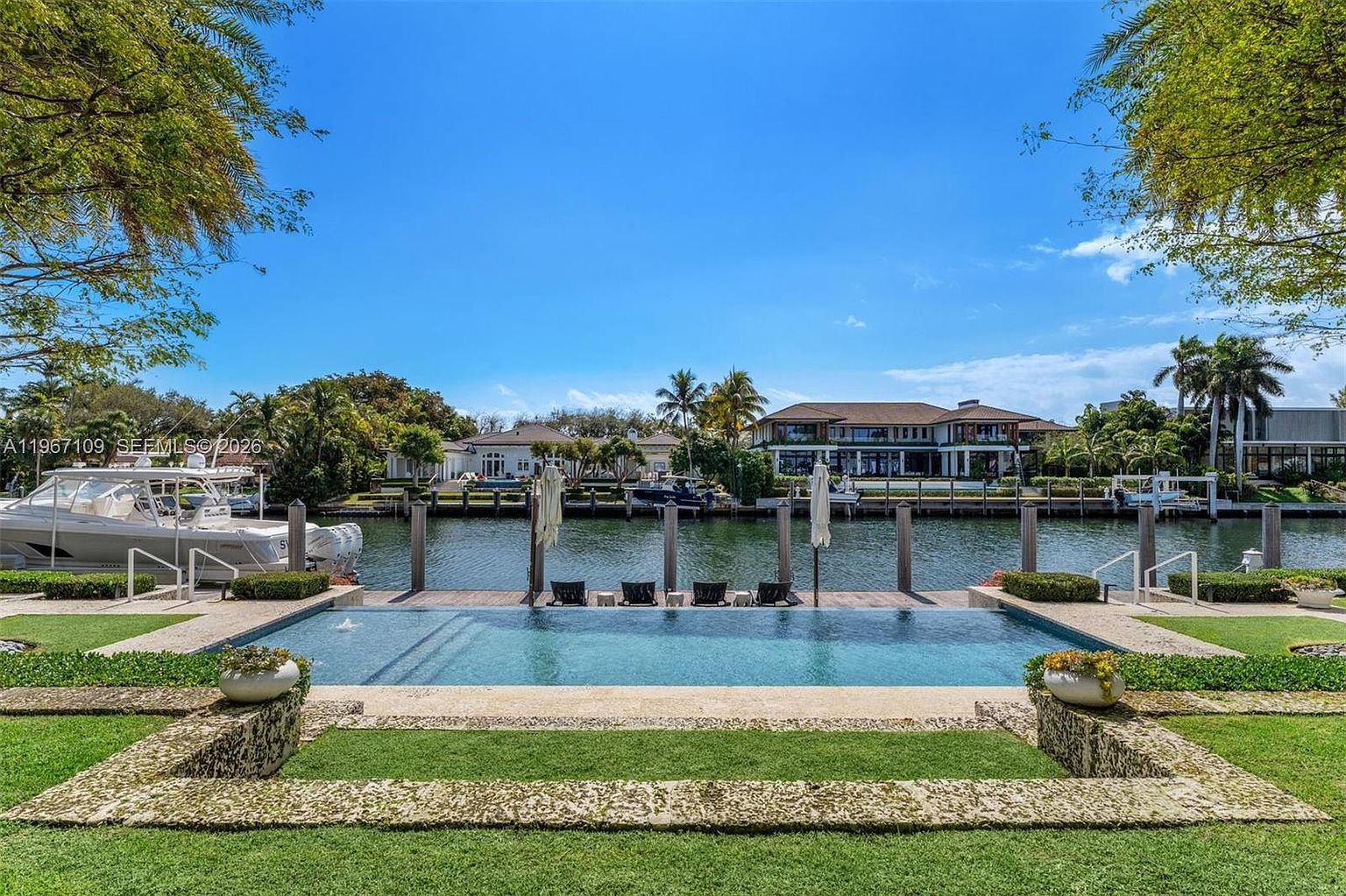 This expansive waterfront property features a sleek, rectangular infinity-edge pool that seamlessly blends into the canal view. The foreground includes a manicured lawn and stone-bordered planters, while the background showcases a private dock with lounge chairs and a large luxury residence across the water. The scene is captured from a low, cinematic perspective, emphasizing the serene connection between the pool, the waterway, and the lush tropical landscaping.