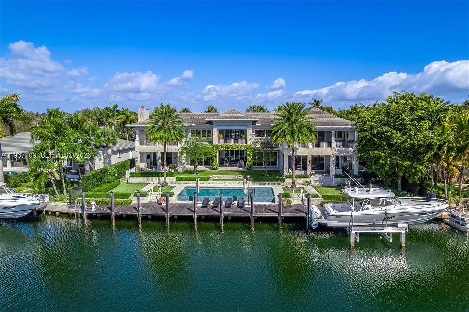 This expansive two-story waterfront estate features a classic architectural design with a symmetrical facade, a tiled roof, and multiple balconies overlooking the water. The property includes a private dock with two boats, a rectangular swimming pool set into a stone patio, and meticulously manicured landscaping. The perspective is an elevated, straight-on shot that captures the entire rear elevation of the home and its integration with the canal.