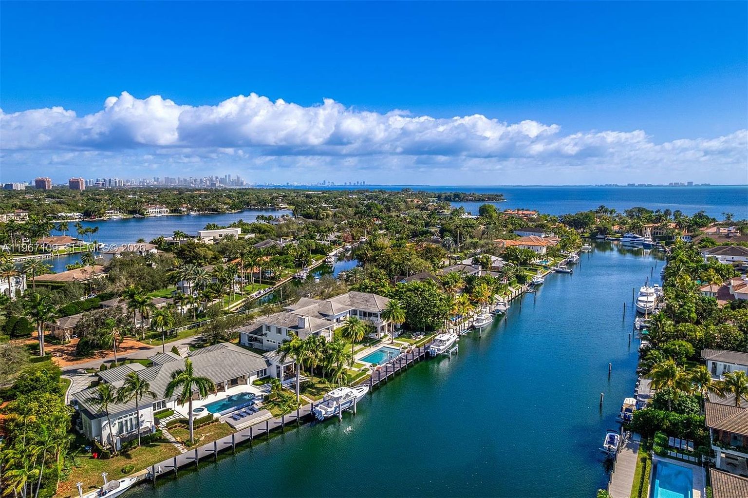 This high-angle aerial view captures a luxurious waterfront residential neighborhood characterized by winding canals and lush tropical landscaping. Several expansive homes with private docks, swimming pools, and manicured lawns line the waterways, showcasing a serene and upscale coastal lifestyle. In the distance, the horizon reveals a clear blue sky and a glimpse of a distant city skyline across the open water.