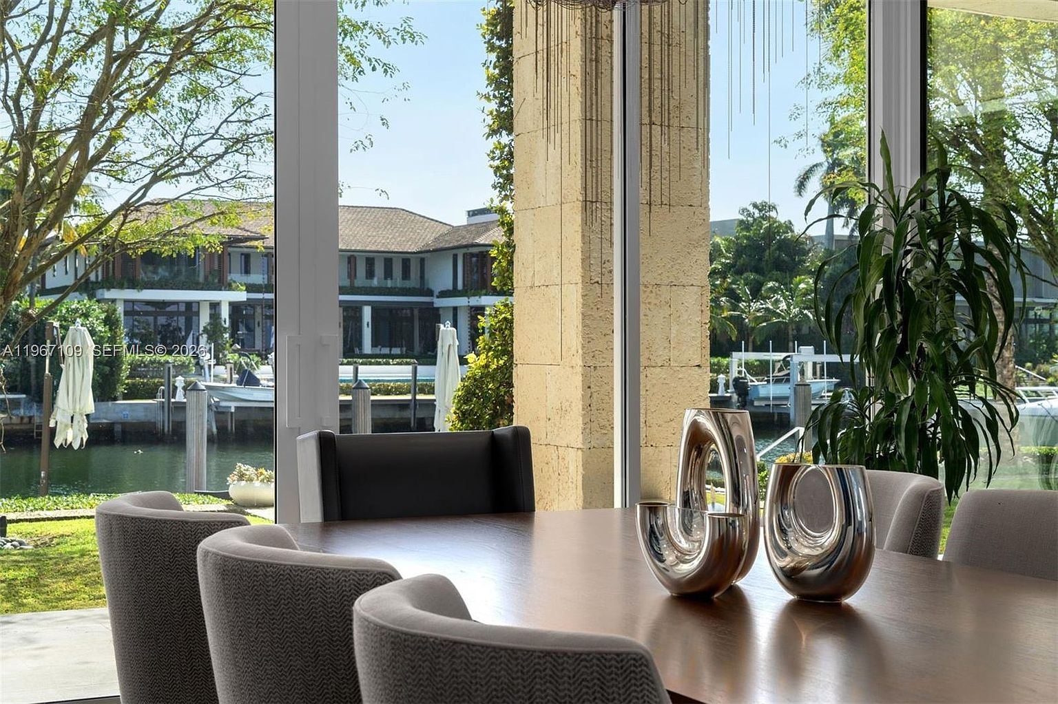 This dining area features a dark wood table surrounded by upholstered chairs, accented by two sculptural silver centerpieces. The space is framed by expansive floor-to-ceiling windows that provide a clear view of a waterfront canal, a boat dock, and neighboring homes. The interior design is complemented by a textured stone pillar, creating a sophisticated and serene atmosphere.