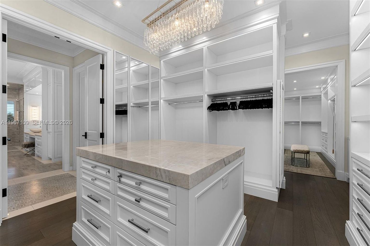 This luxurious walk-in closet features custom white cabinetry with extensive shelving, hanging rods, and a central island topped with a light-toned stone surface. A crystal chandelier hangs above the island, adding a touch of elegance to the space, while dark wood flooring provides a rich contrast to the bright white built-ins. The perspective looks through the closet toward an adjacent bathroom, showcasing a seamless and sophisticated layout.