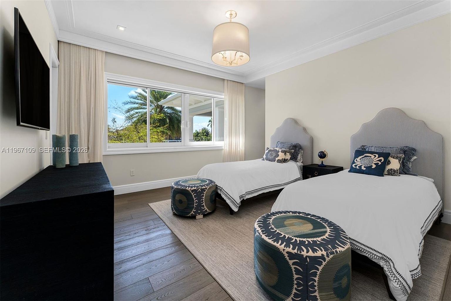 This bright and airy guest bedroom features two twin beds with upholstered headboards and crisp white bedding. A large window allows for ample natural light and a view of the outdoors, while a central drum pendant light provides soft illumination. The room is finished with dark wood flooring, a neutral area rug, and two patterned circular ottomans at the foot of each bed.