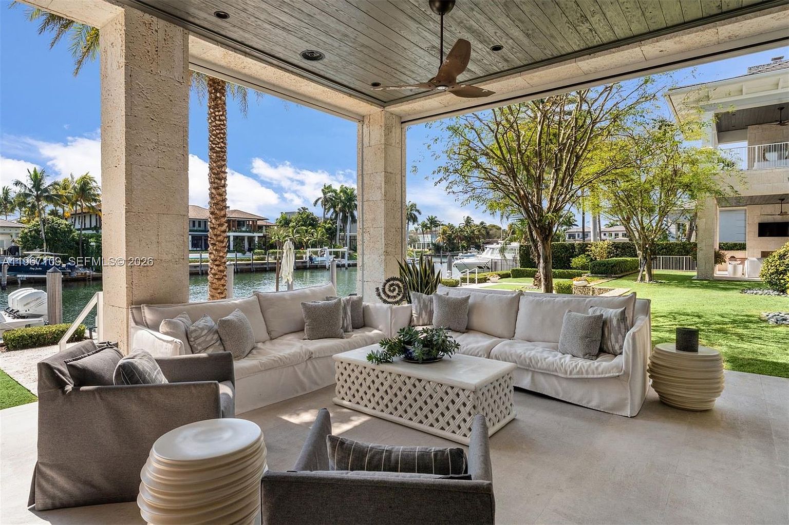 This expansive covered patio offers a luxurious outdoor living space, featuring a large, plush sectional sofa and comfortable armchairs arranged around a decorative white lattice-patterned coffee table. The area is framed by sturdy stone columns and a wood-paneled ceiling with a central fan, providing a seamless transition to the lush green lawn and waterfront view beyond. The setting exudes a serene, resort-like atmosphere, perfect for relaxation and entertaining in a tropical climate.