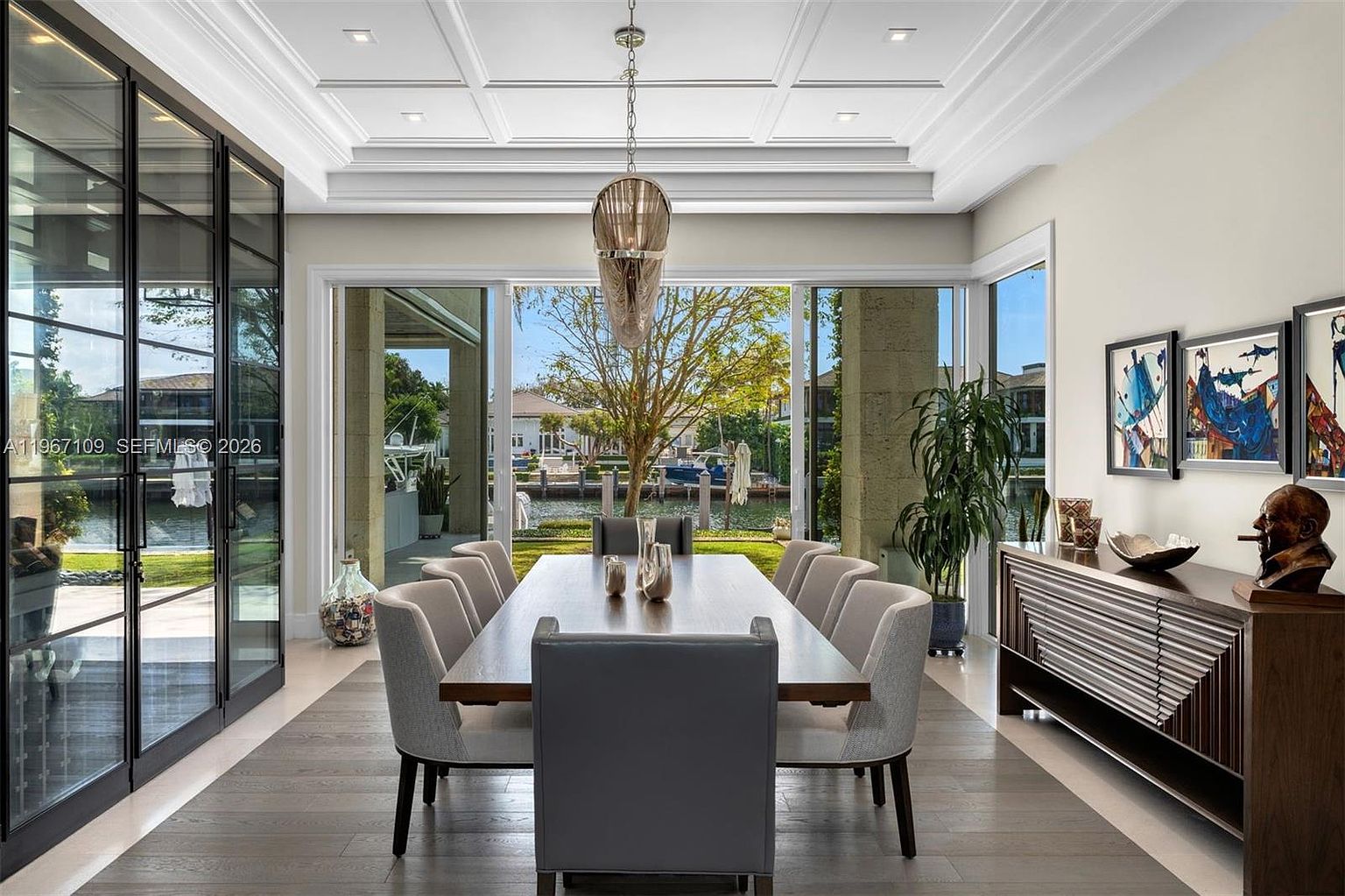 This elegant dining room features a long, dark wood table surrounded by eight upholstered chairs, centered beneath a unique, textured pendant light. The room is framed by large glass sliding doors that provide an expansive view of the waterfront backyard and canal. To the right, a modern sideboard with a geometric wood-paneled front displays decorative art and a bust, creating a sophisticated and inviting atmosphere.