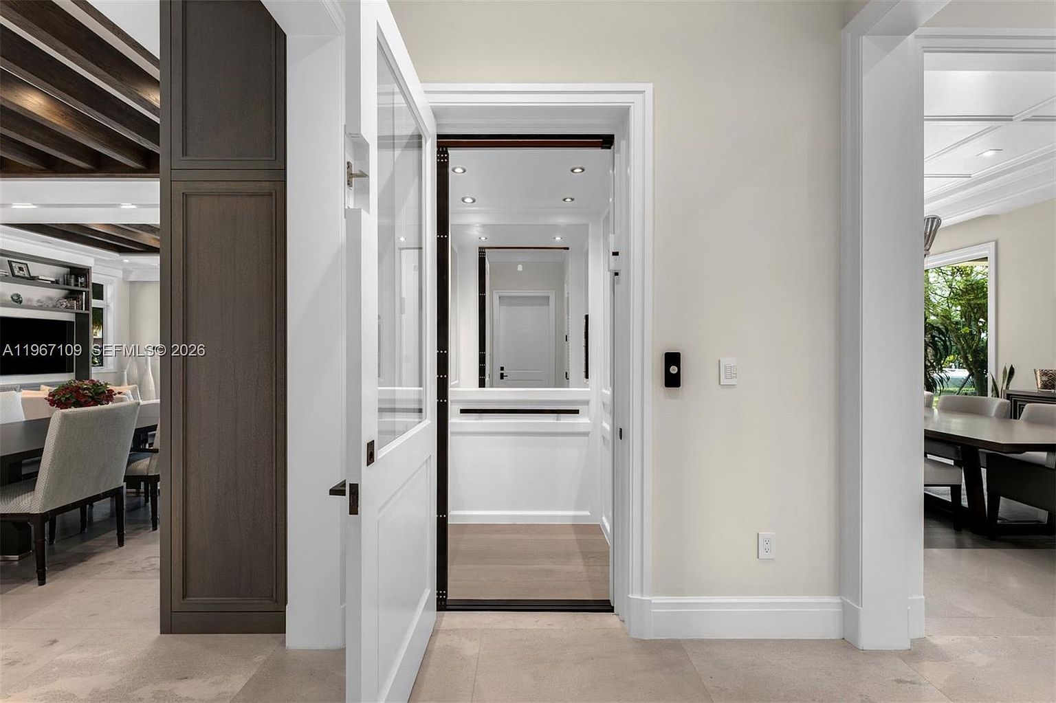 This image captures a residential elevator with an open white door, revealing a mirrored interior that reflects the surrounding hallway. The space features neutral-toned walls, light-colored flooring, and a clean, modern aesthetic that connects to adjacent living and dining areas. The perspective is centered on the elevator entrance, highlighting its integration into the home's layout.